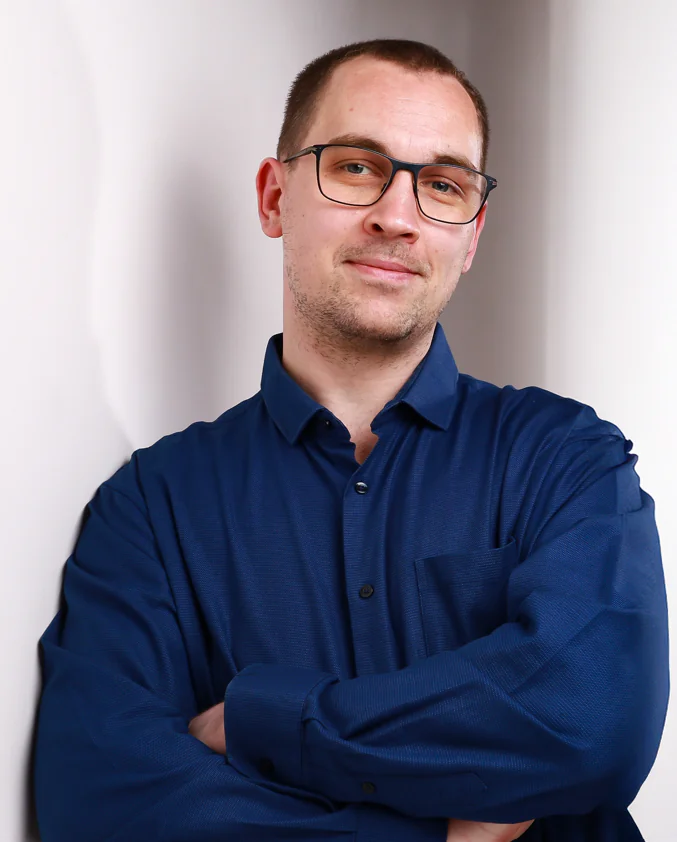 Ein Mann mit Brille, einem dunkelblauen Hemd, die Hände gefaltet, lächelnd.
